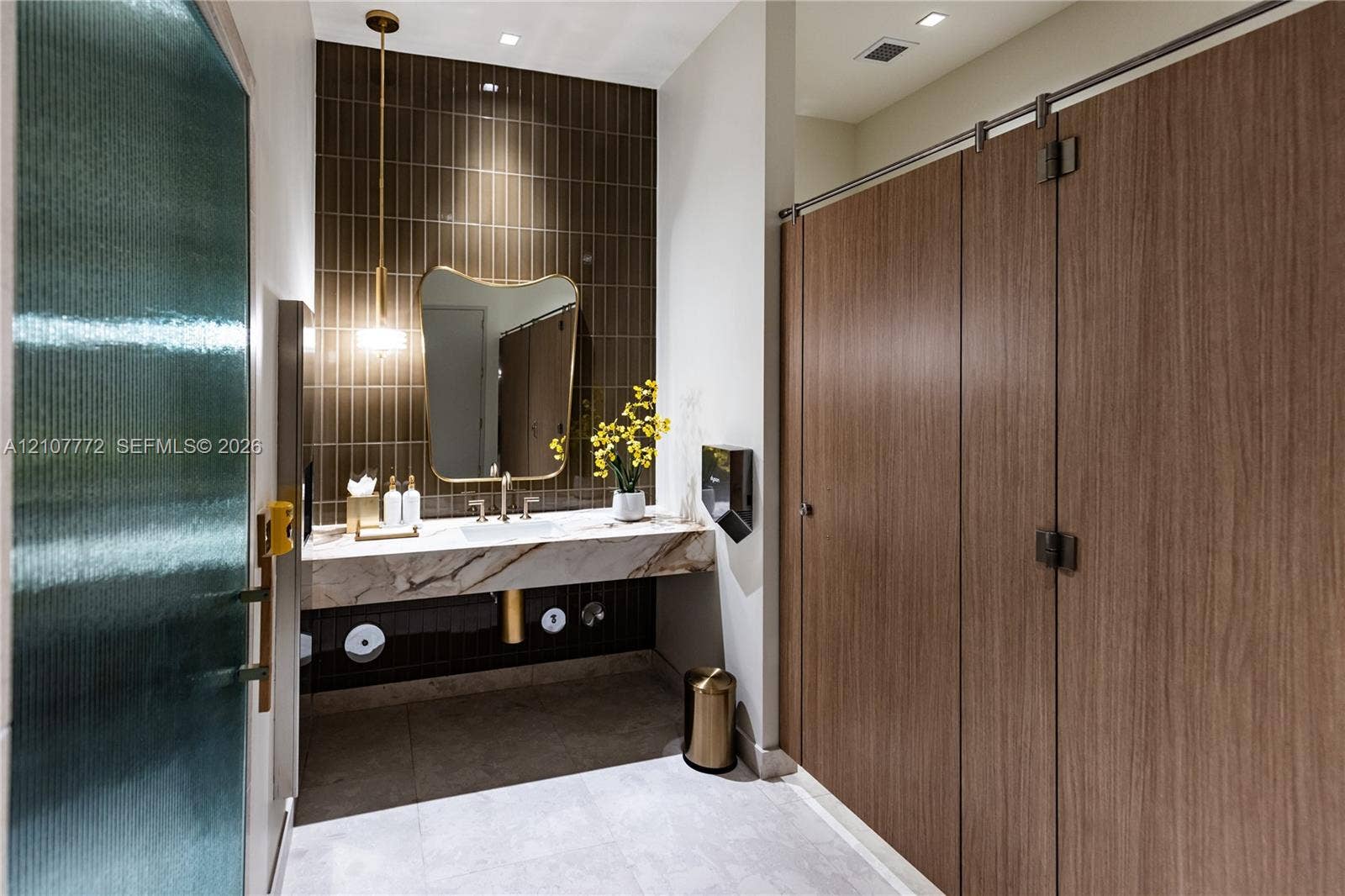 77 SE 5th Street, #1210, Miami, Miami, FL 33131 Floating vanity with a stone countertop, vertical tile backsplash, gold-tone pendant lighting, irregular-shaped mirror, and wood-finish partition doors
