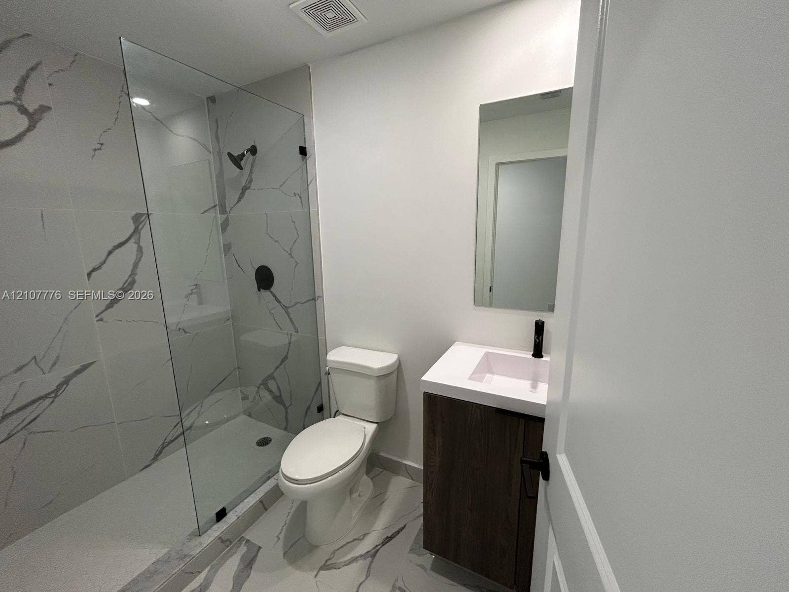 Walk-in shower with floor-to-ceiling veined tile, wood-finish floating vanity with a white countertop, matte black fixtures, and a frameless glass enclosure