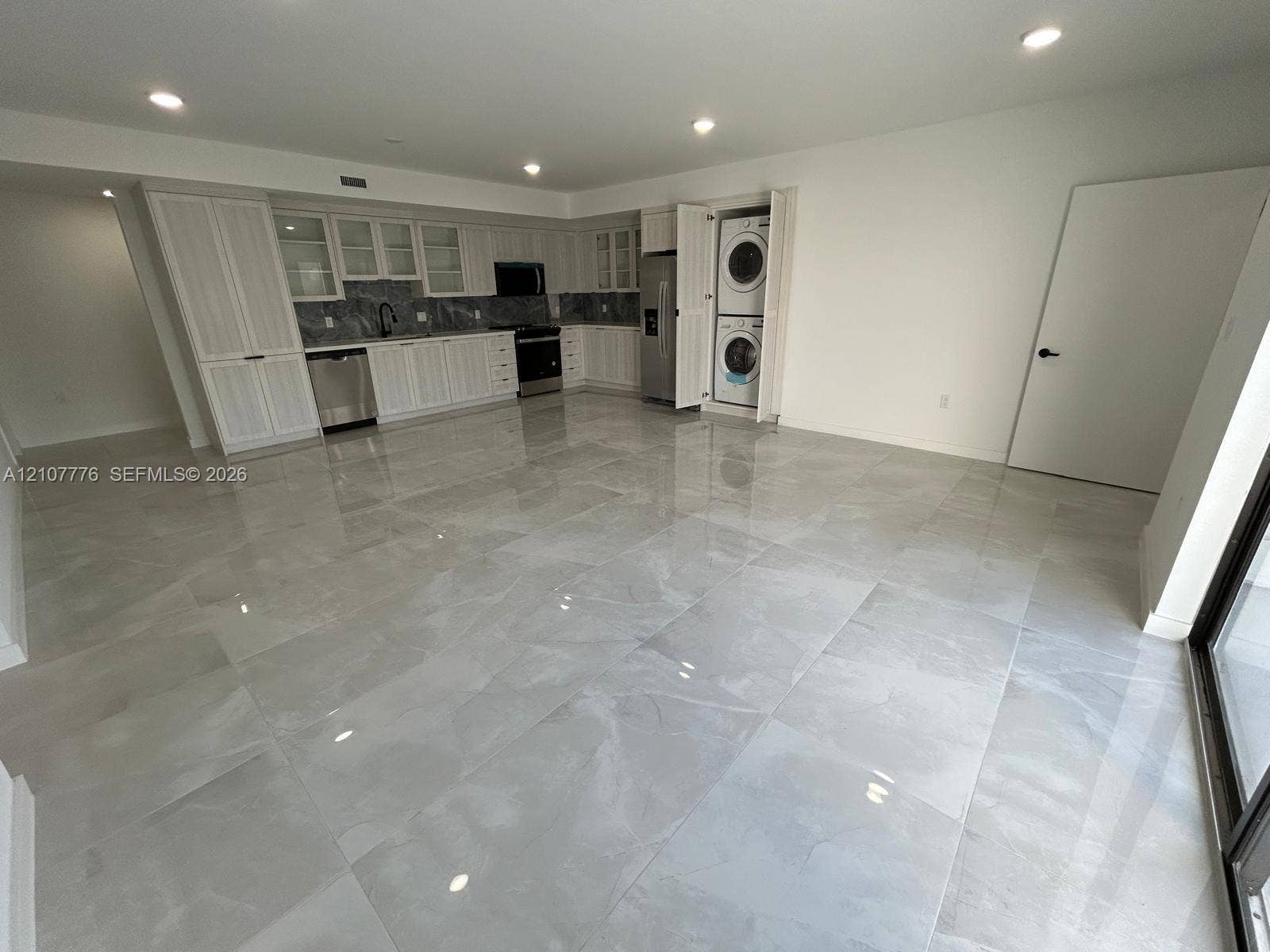 Large-format tile flooring, white cabinetry with integrated upper glass-front displays, stone-finish backsplash, stainless steel appliances, and built-in stacked laundry