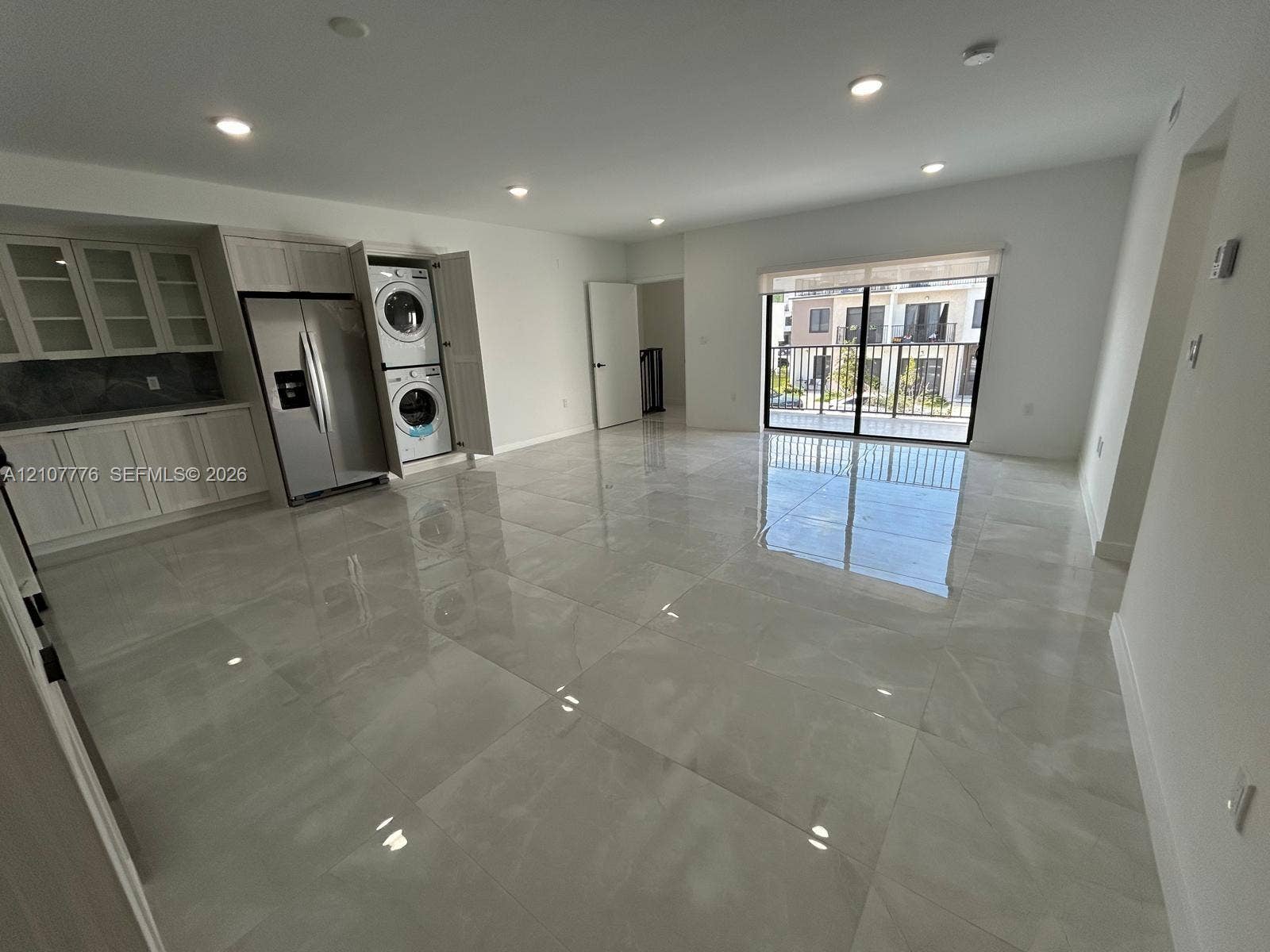 Open-concept interior featuring large-format tile flooring, recessed lighting, white cabinetry with glass-front uppers, stainless steel refrigerator, and a sliding glass door providing external access