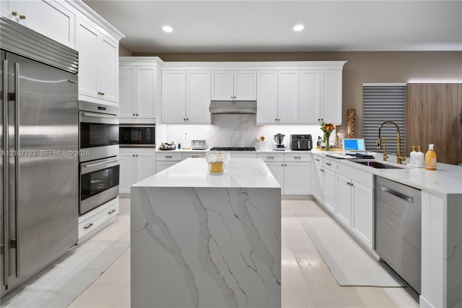 9328 Tropez Lane, Delray Beach, FL 33446 Kitchen featuring white shaker cabinetry, a waterfall-edge center island with stone-finish countertops, stainless steel appliances, a professional-style range hood, and gold-tone faucet hardware