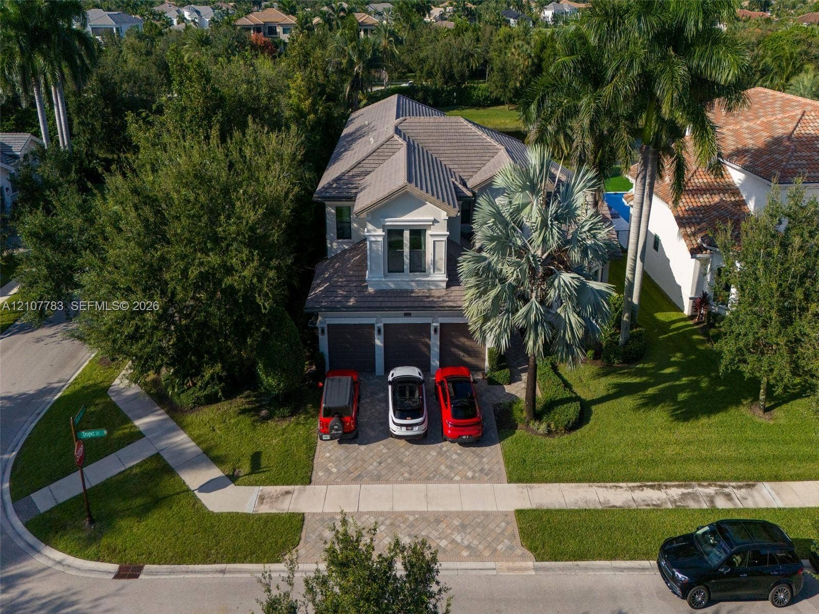 9328 Tropez Lane, Delray Beach, FL 33446 Aerial perspective of a multi-story residence featuring a tile roof, three-car garage, and a paved stone driveway situated on a landscaped lot with mature palm trees
