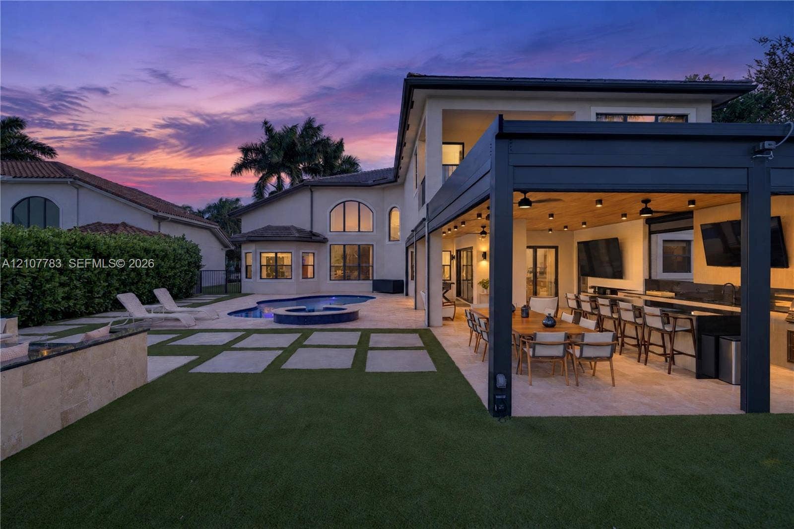 9328 Tropez Lane, Delray Beach, FL 33446 Rear exterior featuring a covered patio with a built-in outdoor kitchen, a swimming pool with an integrated spa, stone-paver hardscaping, and synthetic turf lawn