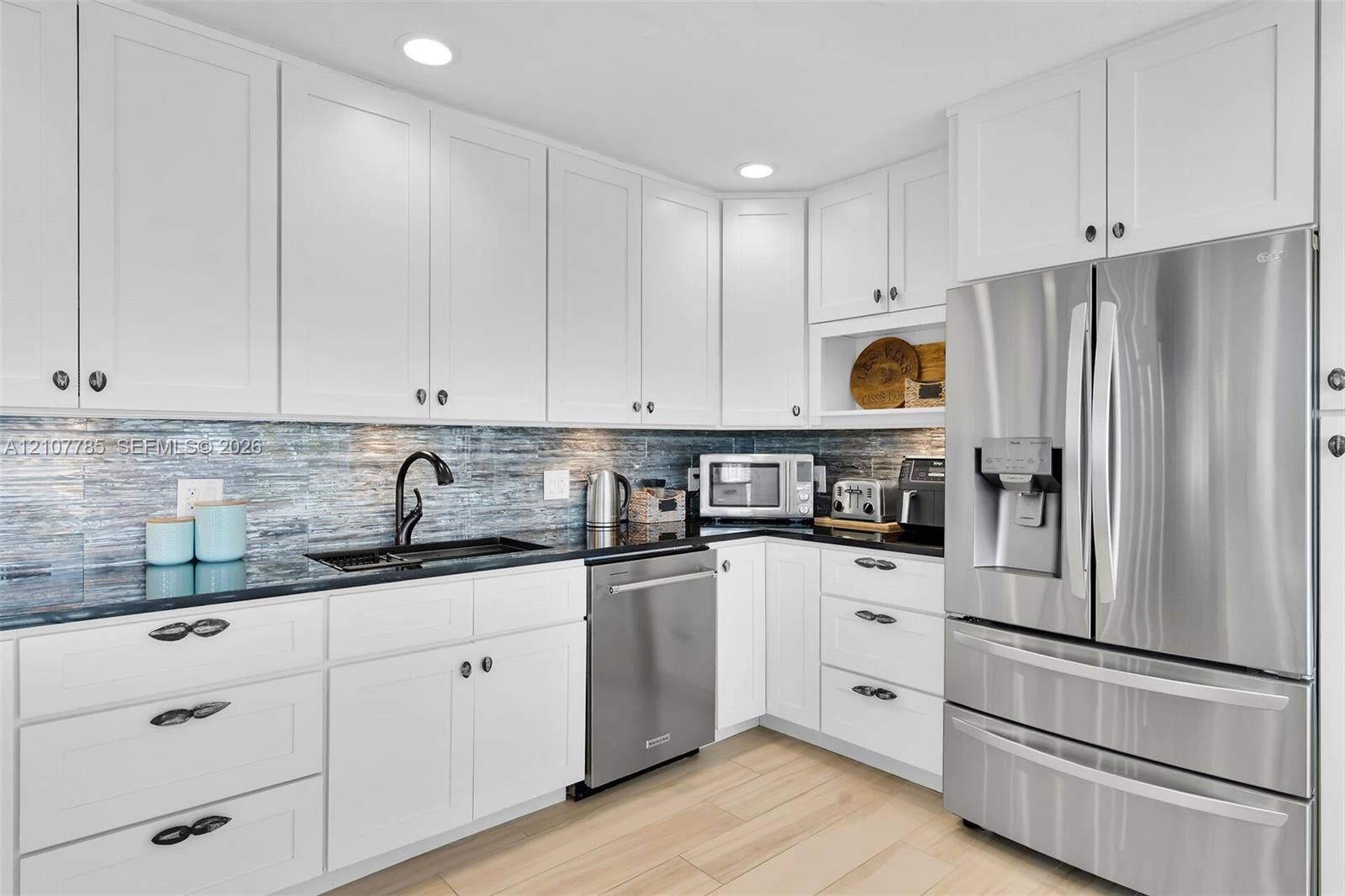Kitchen featuring white shaker-style cabinetry, dark stone countertops, blue glass tile backsplash, stainless steel appliances, and wood-finish flooring