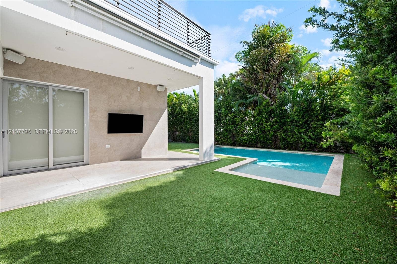 Covered patio featuring stone-tiled walls, swimming pool with integrated sun shelf, synthetic turf lawn, and second-level balcony with metal railing