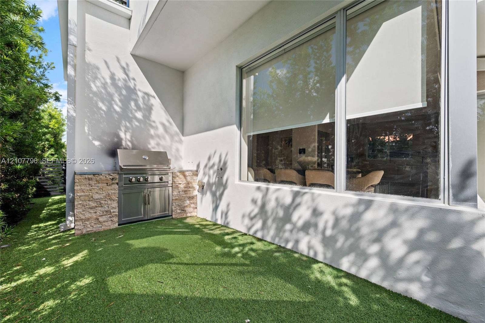 Exterior patio featuring a built-in stainless steel grill, stacked stone facade, large glass windows, and synthetic turf lawn