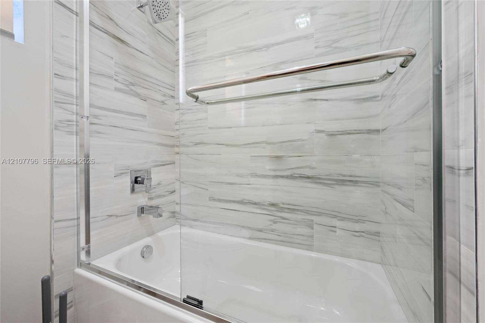 Bathtub featuring floor-to-ceiling stone-look tile surround, glass pivot door, wall-mounted chrome showerhead, and integrated chrome towel bar
