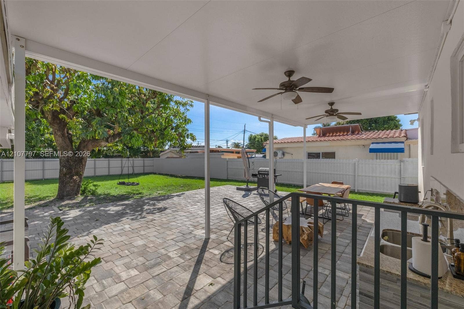 Covered patio featuring a paved stone surface, dual ceiling fans, built-in outdoor kitchen with stone-finish cabinetry, and a vinyl perimeter fence enclosing the lawn