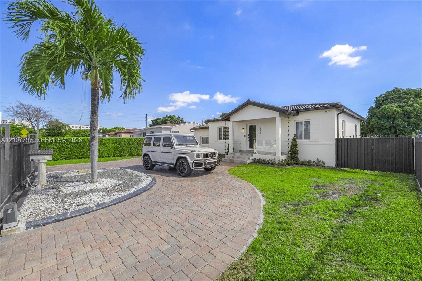 Single-story exterior featuring a paver driveway, covered front entryway, white stucco facade, perimeter fencing, and established palm tree landscaping