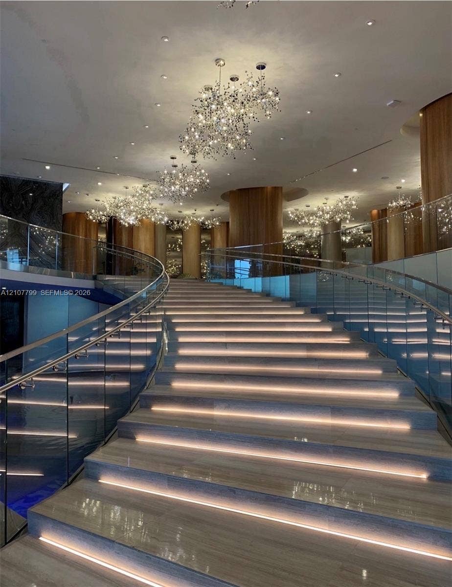 Grand curved staircase featuring stone treads with integrated riser lighting, glass panel railings, polished stone surfaces, and multiple statement crystal chandeliers