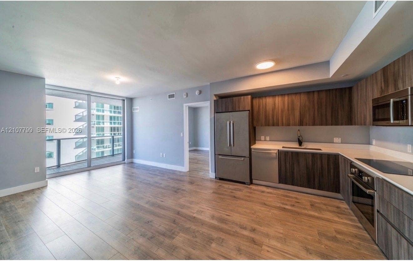 Open-concept kitchen featuring dark wood-finish cabinetry, solid surface countertops, stainless steel appliances, and wood-finish flooring with floor-to-ceiling glass paneling