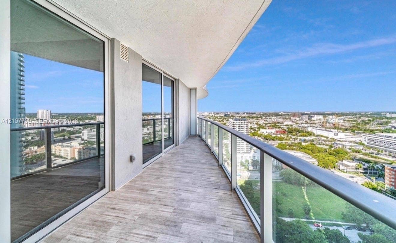 Expansive private balcony featuring wood-finish tile flooring, floor-to-ceiling glass sliding doors, and a glass-paneled railing