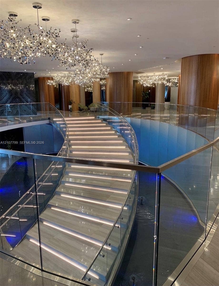 Curved stone staircase with integrated LED tread lighting, glass railings, wood-paneled columns, water feature, and decorative multi-light chandeliers