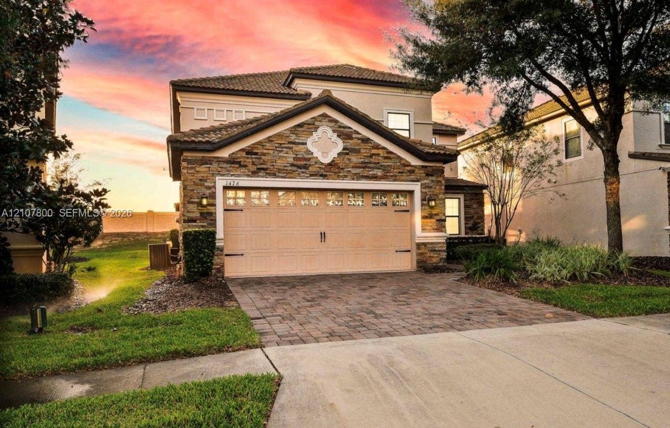 1476 Moon Valley, Davenport, FL 33896 Two-story residence featuring stacked stone veneer, a multi-paneled garage door, decorative gable medallion, tile roofing, and a paved stone driveway