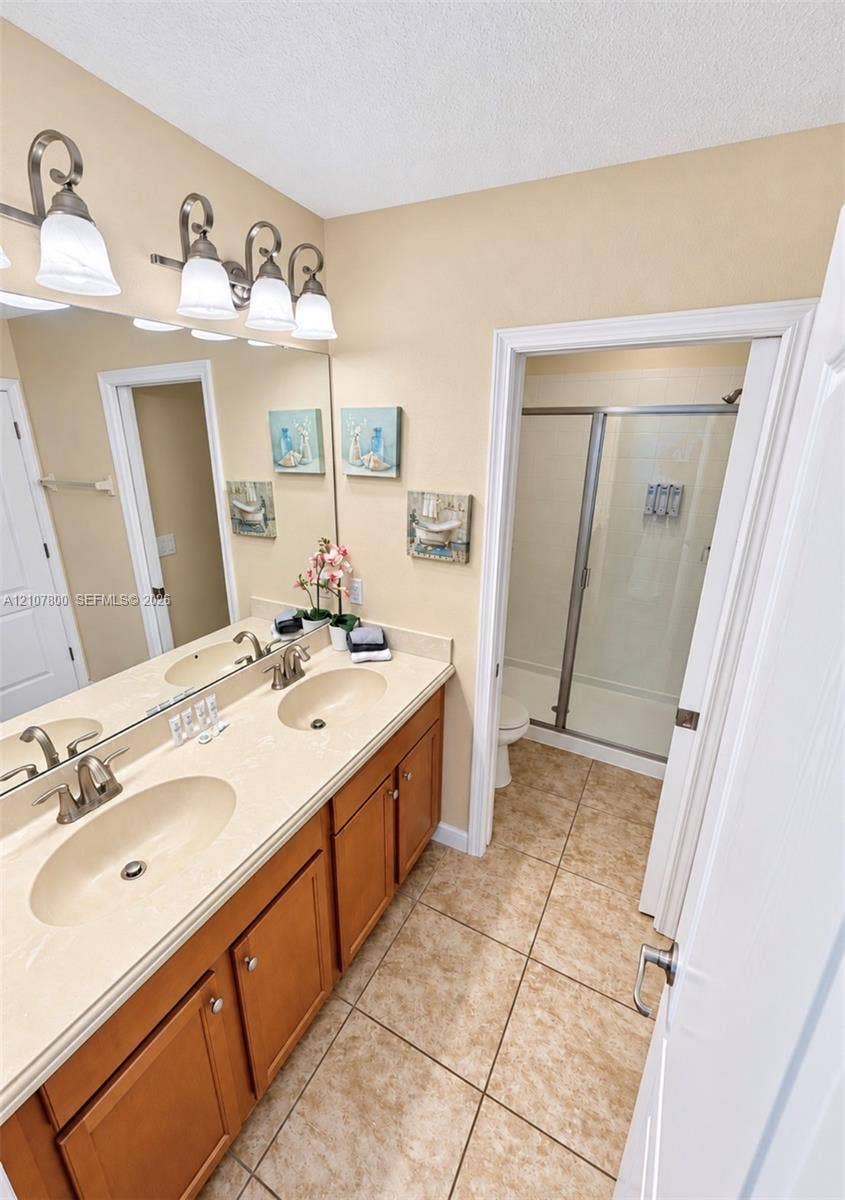 1476 Moon Valley, Davenport, FL 33896 Dual-sink vanity with a solid-surface countertop, four-light vanity fixture, expansive wall-to-wall mirror, tiled flooring, and a walk-in shower with a glass door