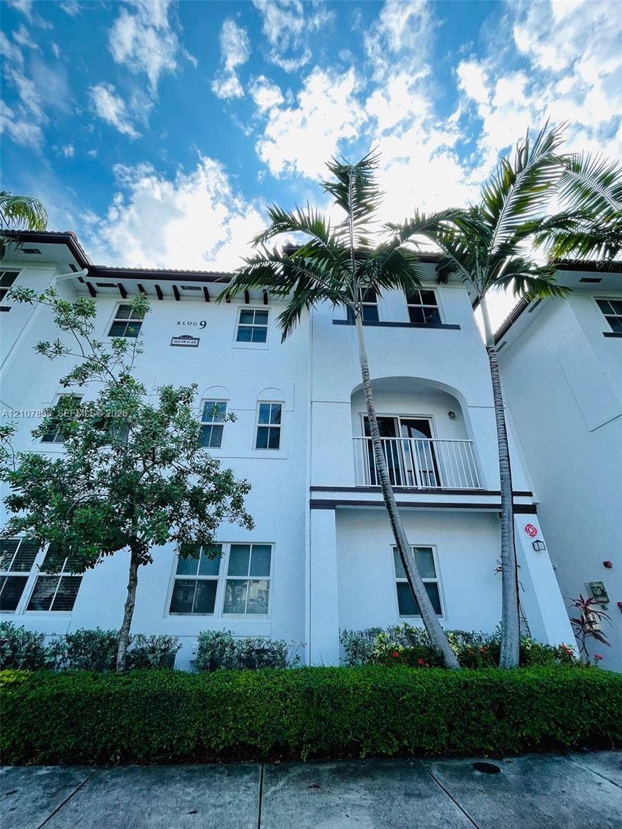 8620 NW 97th Avenue, #205, Doral, FL 33178 Multi-story building facade featuring white stucco finish, decorative bracket eaves, a private balcony with metal railing, and mature palm landscaping