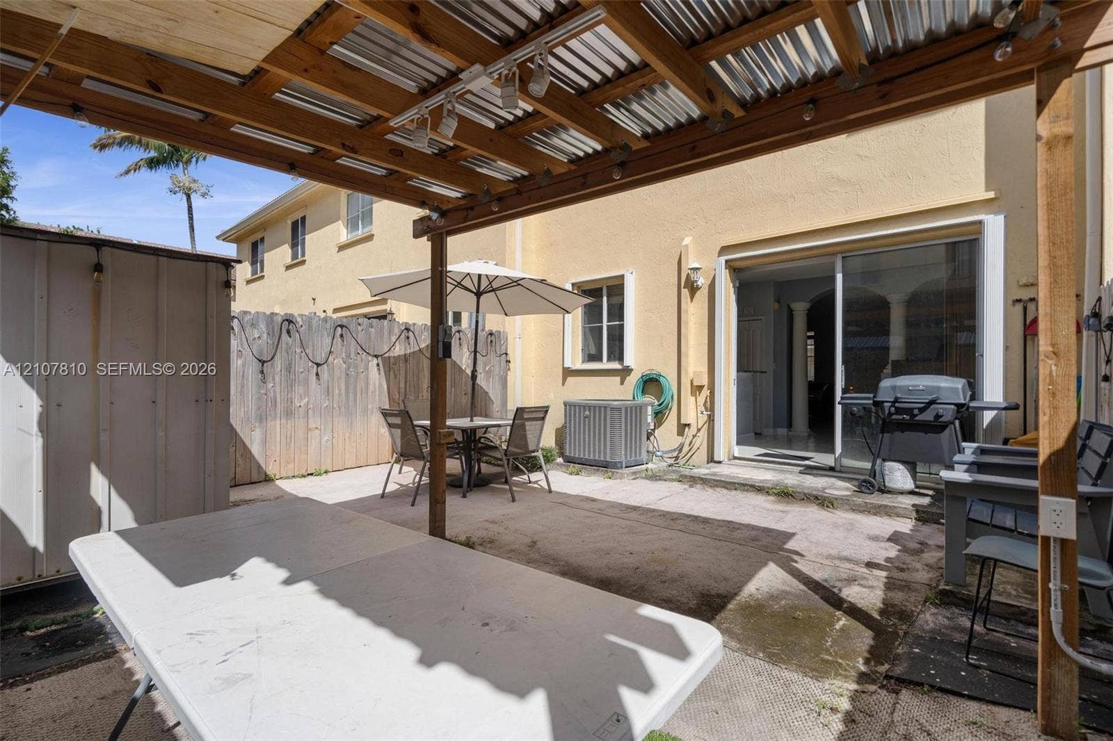 6940 NW 177th Street, #P105, Hialeah, Hialeah, FL 33015 Paved rear patio featuring a corrugated metal and wood-beam pergola, wood privacy fencing, and sliding glass door access. Outdoor furniture, BBQ Grill and Outside TV stays.
