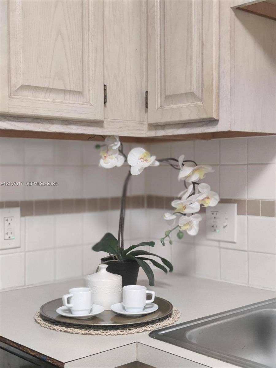 1705 Palm Cove Boulevard, #1-104, Delray Beach, Delray Beach, FL 33445 Light-tone wood cabinetry featuring recessed-panel doors, neutral solid-surface countertops, and square ceramic tile backsplash with decorative accent inlay