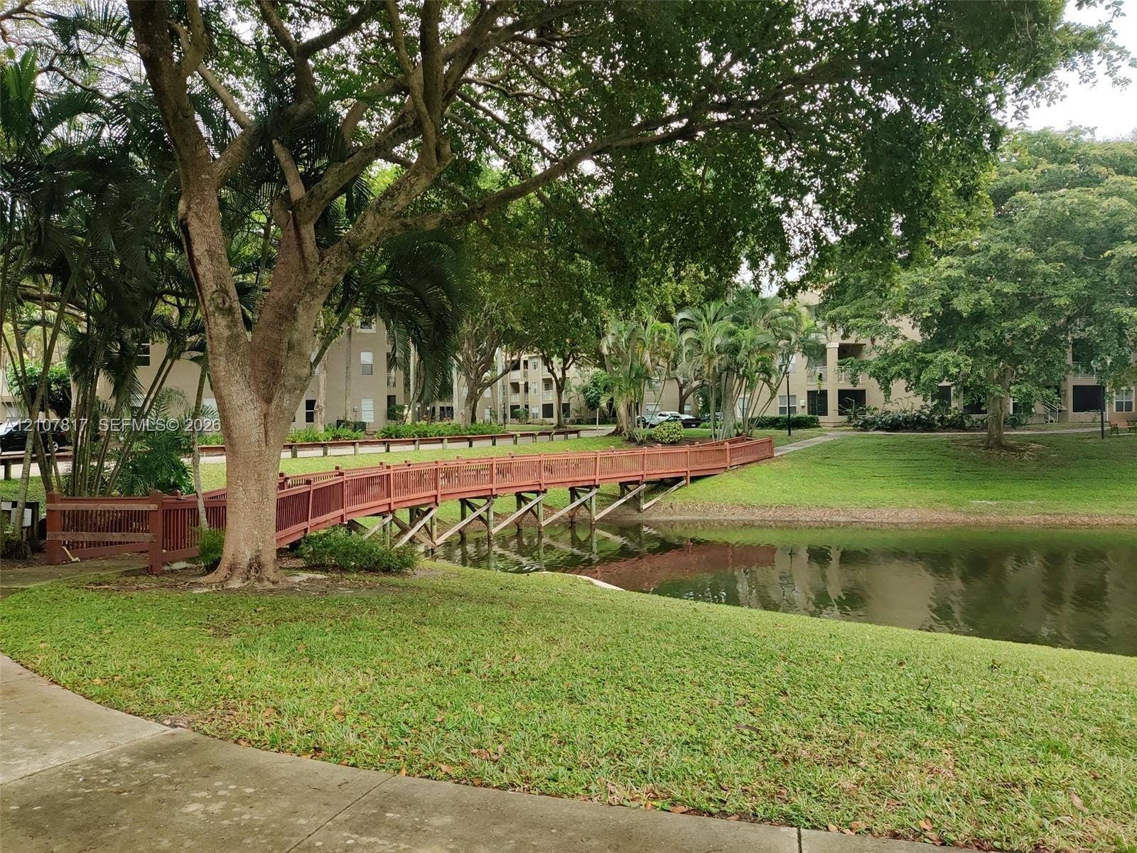 1705 Palm Cove Boulevard, #1-104, Delray Beach, Delray Beach, FL 33445 Waterfront setting featuring a wooden pedestrian bridge, manicured landscaping, mature trees, and concrete paved pathways