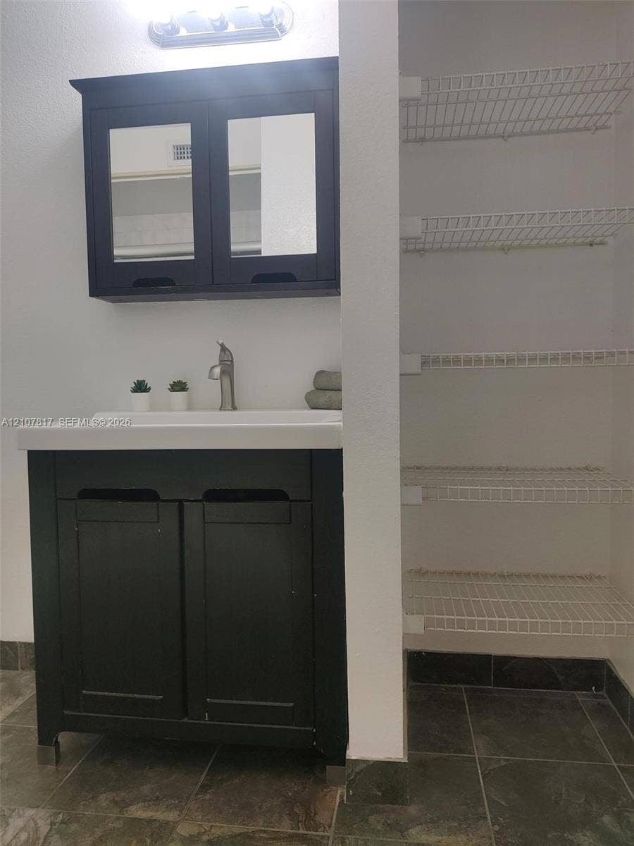 1705 Palm Cove Boulevard, #1-104, Delray Beach, Delray Beach, FL 33445 Dark-finish vanity with integrated rectangular sink and matching mirrored cabinet, accompanied by a multi-level wire shelving unit and tile flooring