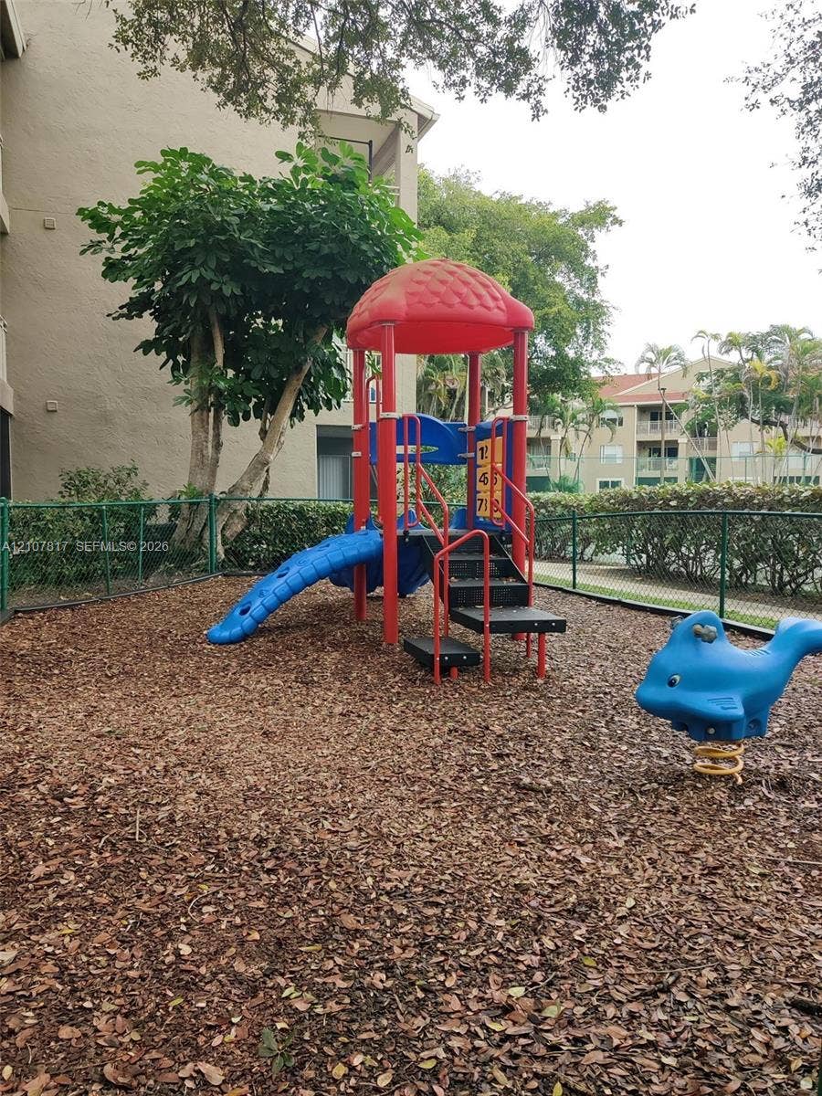 1705 Palm Cove Boulevard, #1-104, Delray Beach, Delray Beach, FL 33445 Outdoor recreational area featuring a multi-level climbing structure with slide, spring-mounted animal rider, wood chip surfacing, and perimeter chain-link fencing
