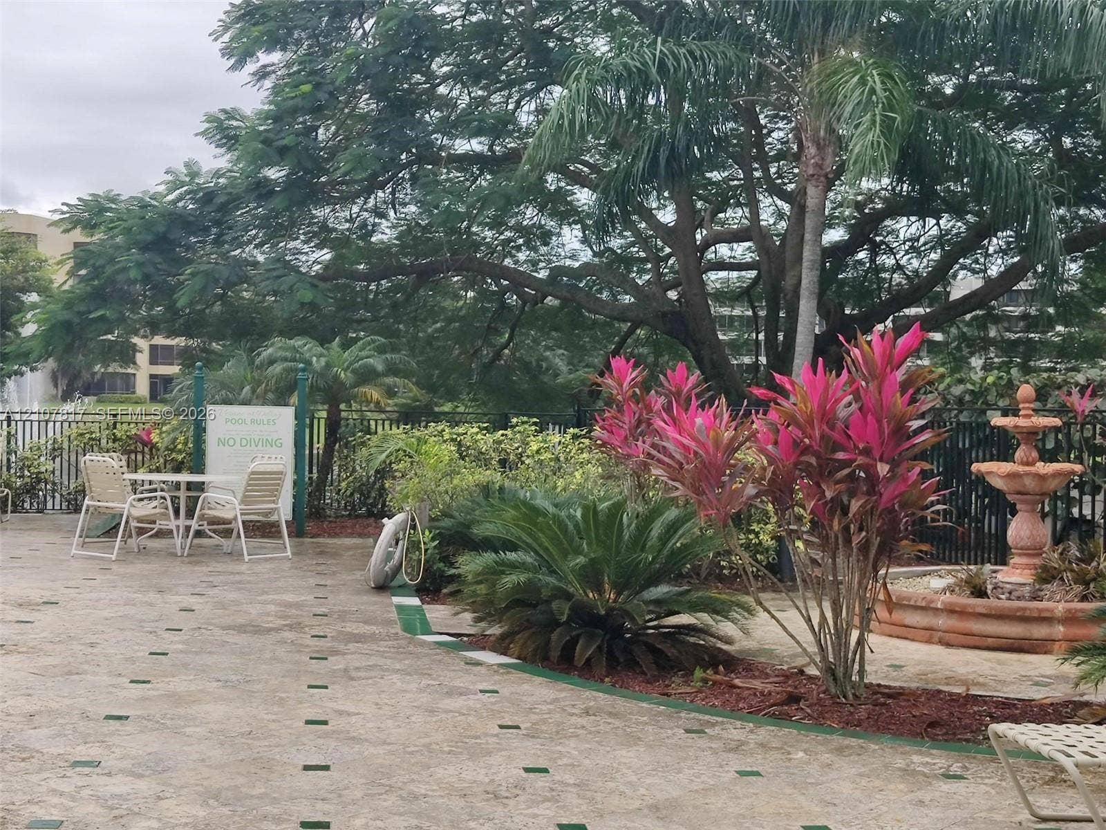 1705 Palm Cove Boulevard, #1-104, Delray Beach, Delray Beach, FL 33445 Outdoor patio featuring stone-tile flooring with decorative inlays, a tiered stone fountain, mature tropical landscaping, and metal perimeter fencing