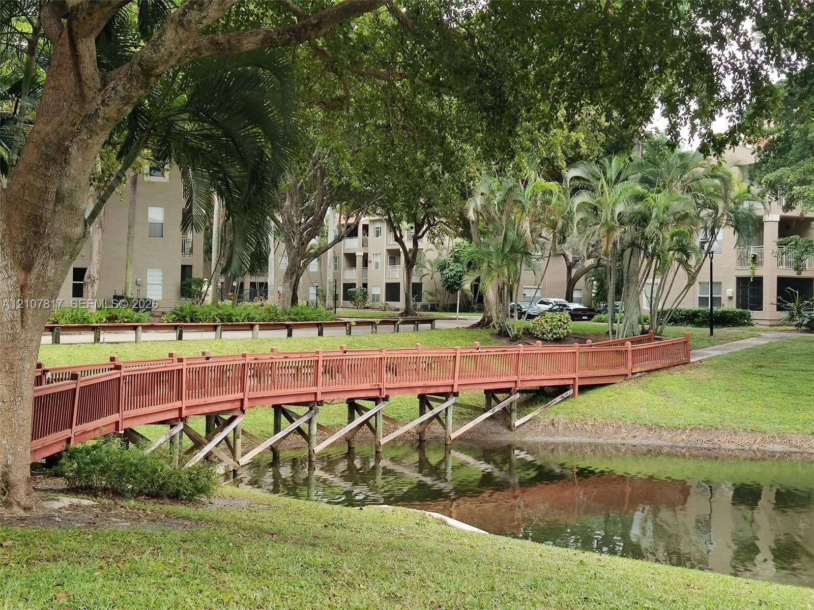 1705 Palm Cove Boulevard, #1-104, Delray Beach, Delray Beach, FL 33445 Pedestrian bridge with wooden railing over a landscaped pond, surrounded by mature trees and manicured green space