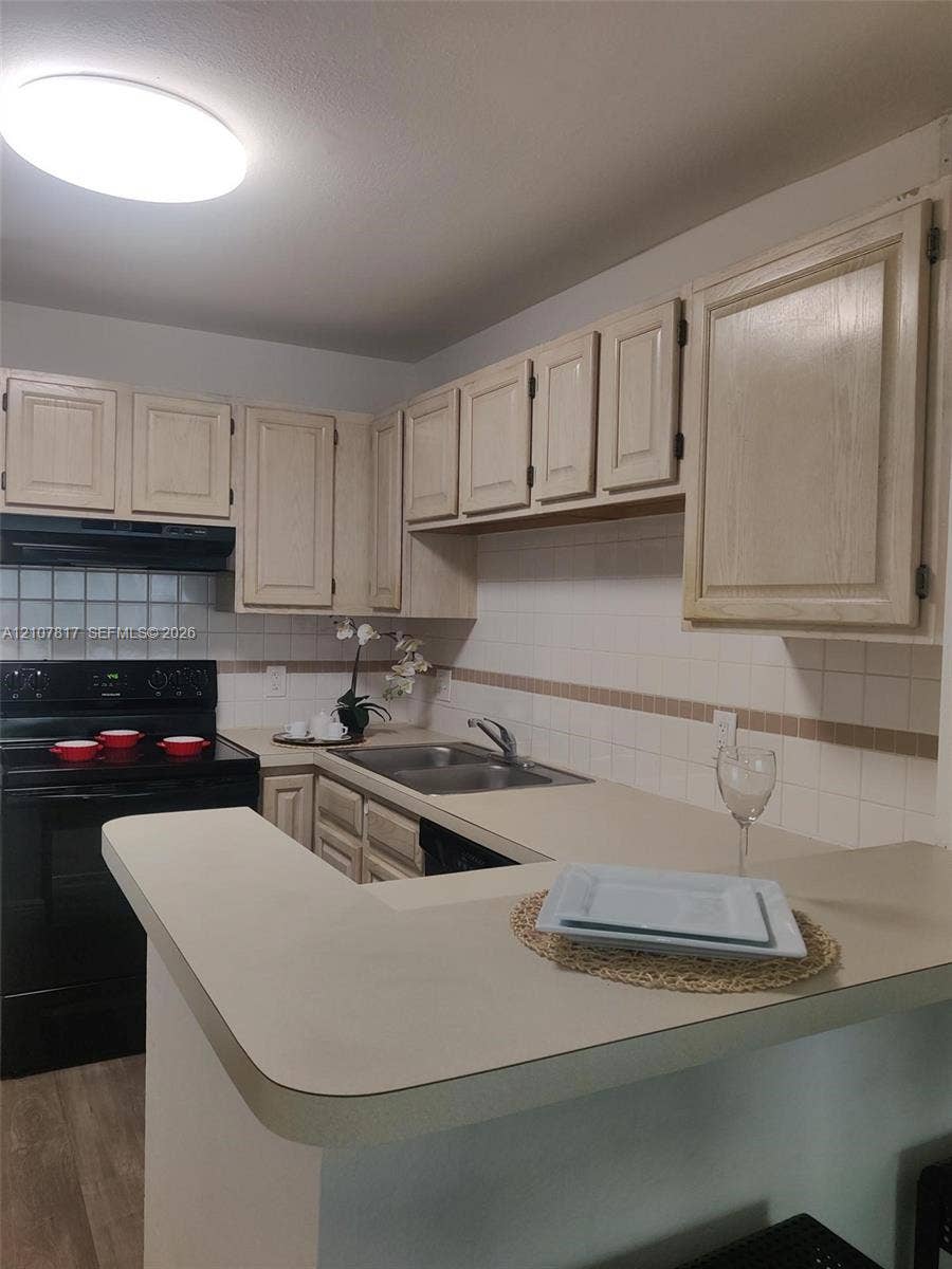 1705 Palm Cove Boulevard, #1-104, Delray Beach, Delray Beach, FL 33445 Kitchen featuring light-toned cabinetry, dual-basin stainless steel sink, solid surface countertop with extended breakfast bar, tiled backsplash, and black appliances