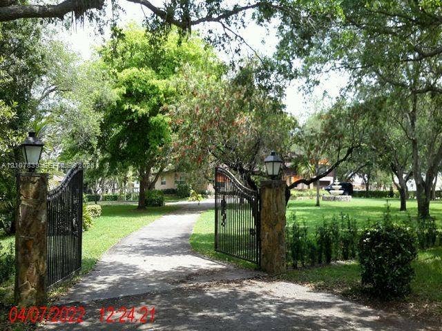 12105 NW 23rd Court, Plantation, FL 33323 Gated entrance featuring stone masonry pillars, wrought iron fencing, mounted exterior lighting fixtures, and a landscaped grounds area with mature trees and a fountain