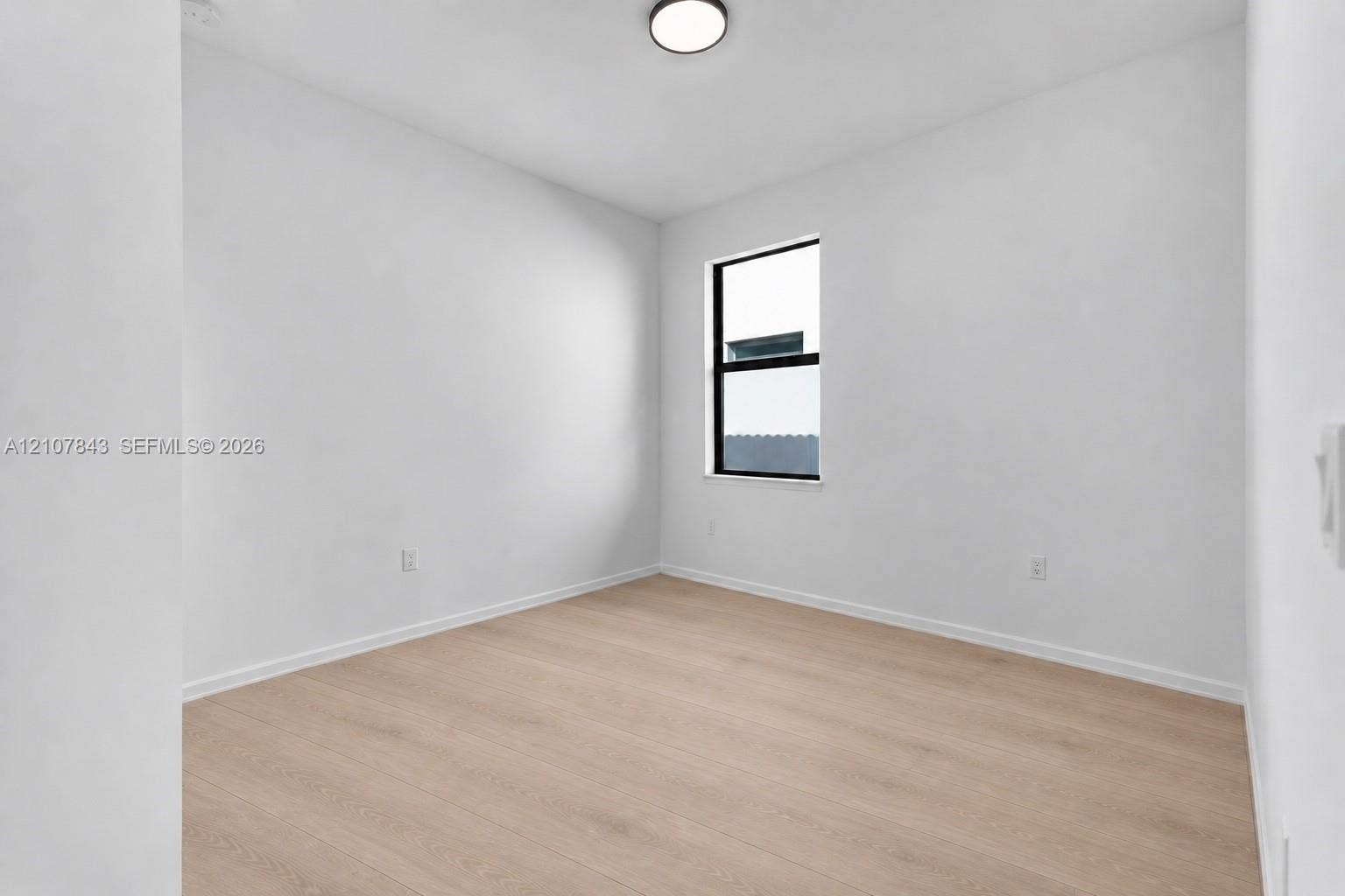 4949 Blistering Way, Lake Worth, FL 33467 Neutral-toned interior space featuring wood-finish flooring, crisp white wall finishes, and a single-pane window with a black frame, completed by a minimalist flush-mount ceiling light fixture