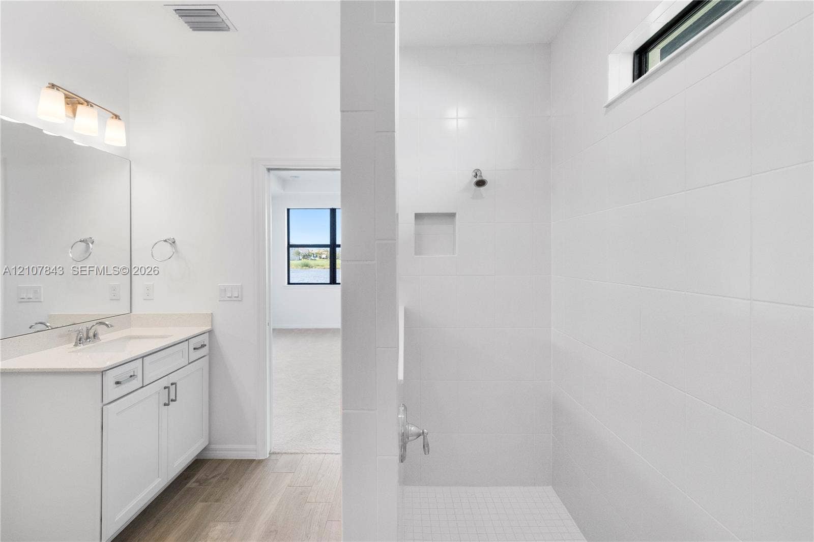 4949 Blistering Way, Lake Worth, FL 33467 Bathroom featuring a white cabinetry vanity with a solid surface countertop, an integrated wall-to-wall mirror with attached light fixture, wood-finish flooring, and a walk-in shower with white wall tile, built-in shelving niche, and transom window