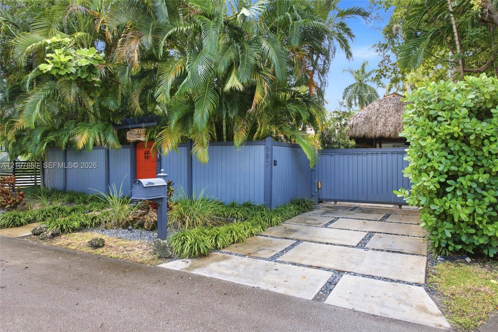Private gated entry featuring a custom blue vertical-board fence, concrete paver driveway with crushed stone detailing, and mature tropical landscaping