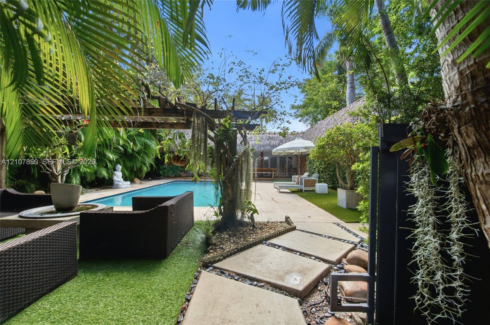 Outdoor swimming pool featuring a wooden pergola, concrete paver walkways, synthetic turf landscaping, and mature tropical greenery