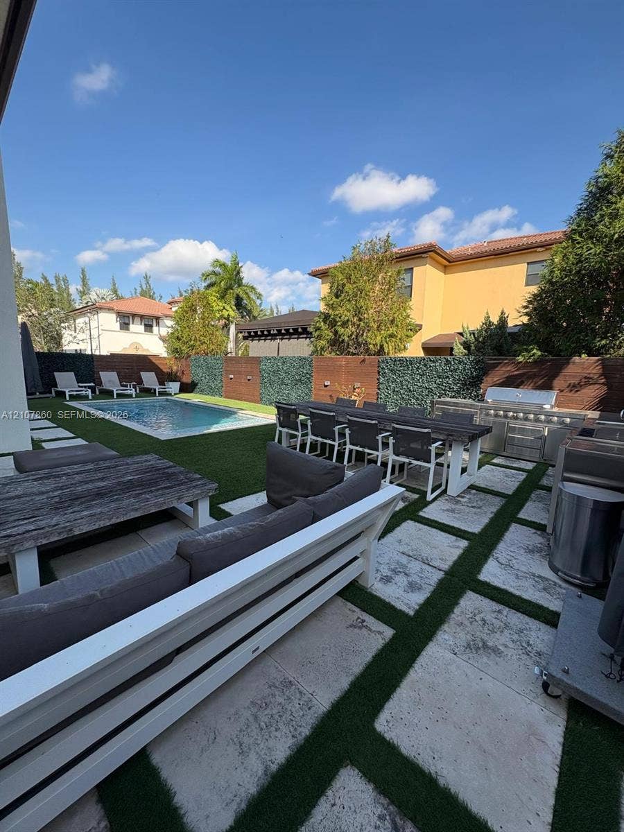 8925 NW 99th Court, Doral, FL 33178 Rear patio featuring natural stone pavers, a built-in stainless steel outdoor kitchen, synthetic lawn, and a rectangular swimming pool enclosed by a privacy wall