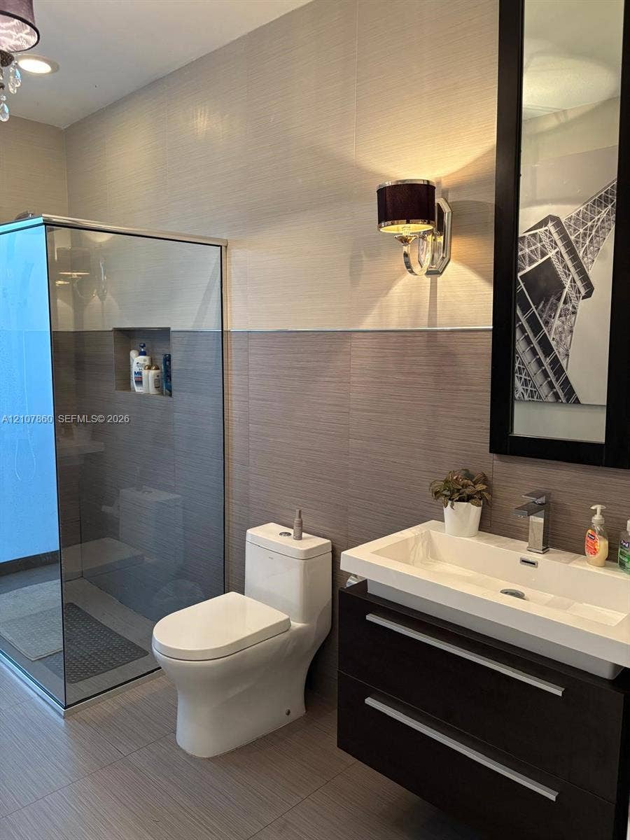8925 NW 99th Court, Doral, FL 33178 Floating vanity with rectangular vessel sink and chrome faucet, glass-enclosed shower featuring a recessed wall niche, two-tone textured wall tile, and wall-mounted drum shade sconce