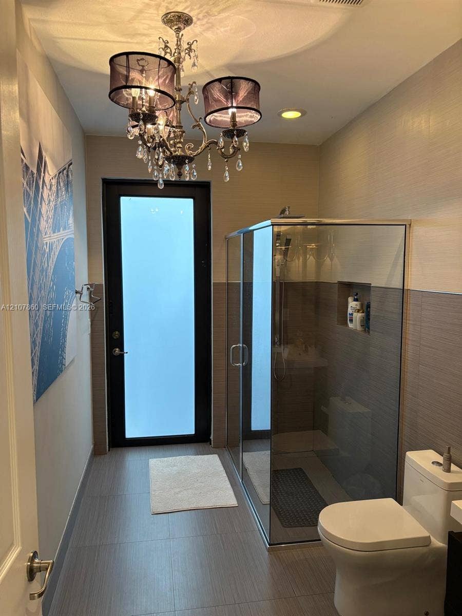 8925 NW 99th Court, Doral, FL 33178 Bathroom featuring a walk-in glass-enclosed shower with a recessed wall niche, dual-tone tiled wall finish, frosted-glass exterior door, and ornate crystal chandelier lighting