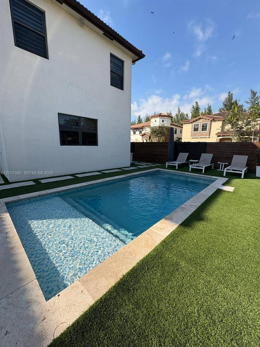 8925 NW 99th Court, Doral, FL 33178 Rectangular swimming pool featuring a tanning ledge and natural stone coping, surrounded by synthetic turf and oversized concrete pavers, enclosed by a horizontal wood fence