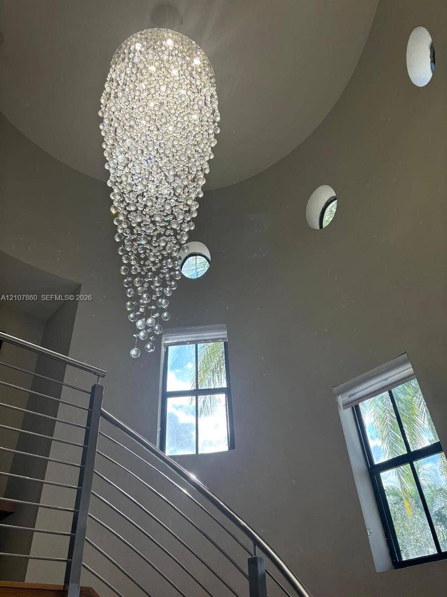 8925 NW 99th Court, Doral, FL 33178 Tapered multi-light crystal chandelier suspended from a vaulted ceiling, featuring circular and rectangular windows and a metal-railing staircase
