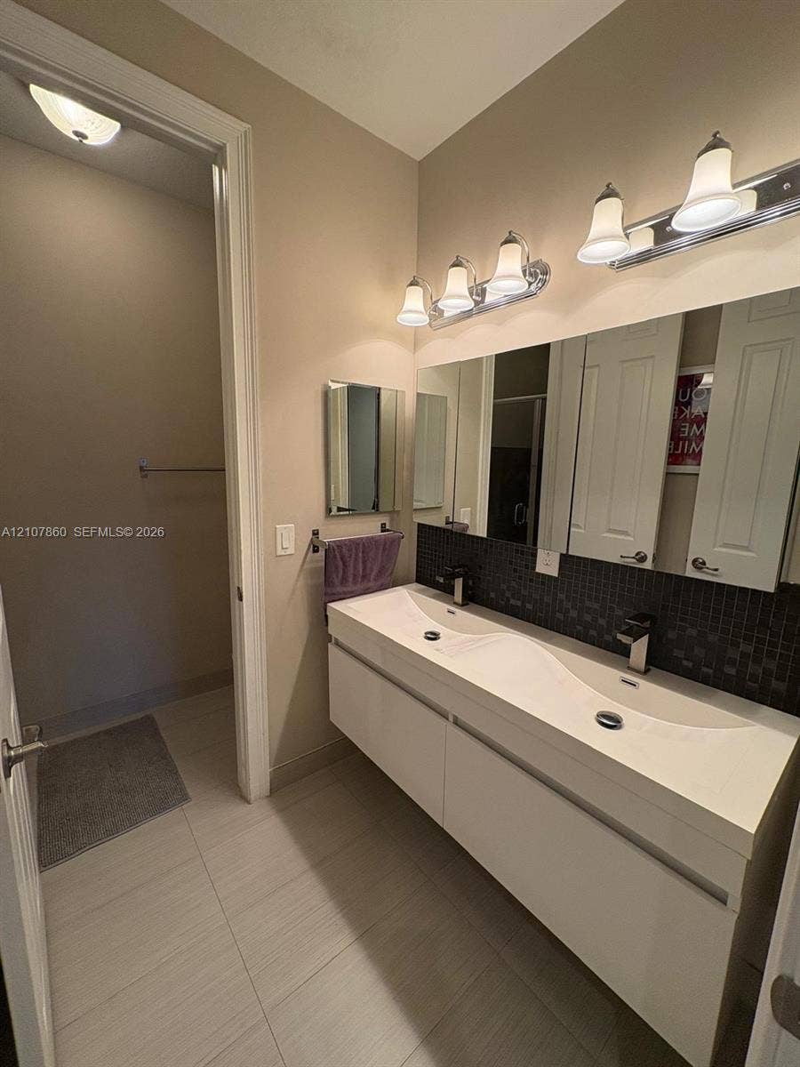 8925 NW 99th Court, Doral, FL 33178 Floating dual-sink vanity with a seamless integrated basin, dark mosaic tile backsplash, large wall-mounted mirror, and multi-bulb polished chrome vanity lighting
