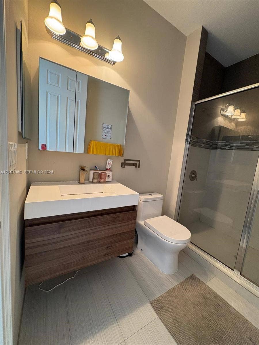 8925 NW 99th Court, Doral, FL 33178 Modern floating wood-finish vanity with integrated sink, three-light vanity fixture, dual-panel medicine cabinet, and glass-enclosed shower with tiled surround
