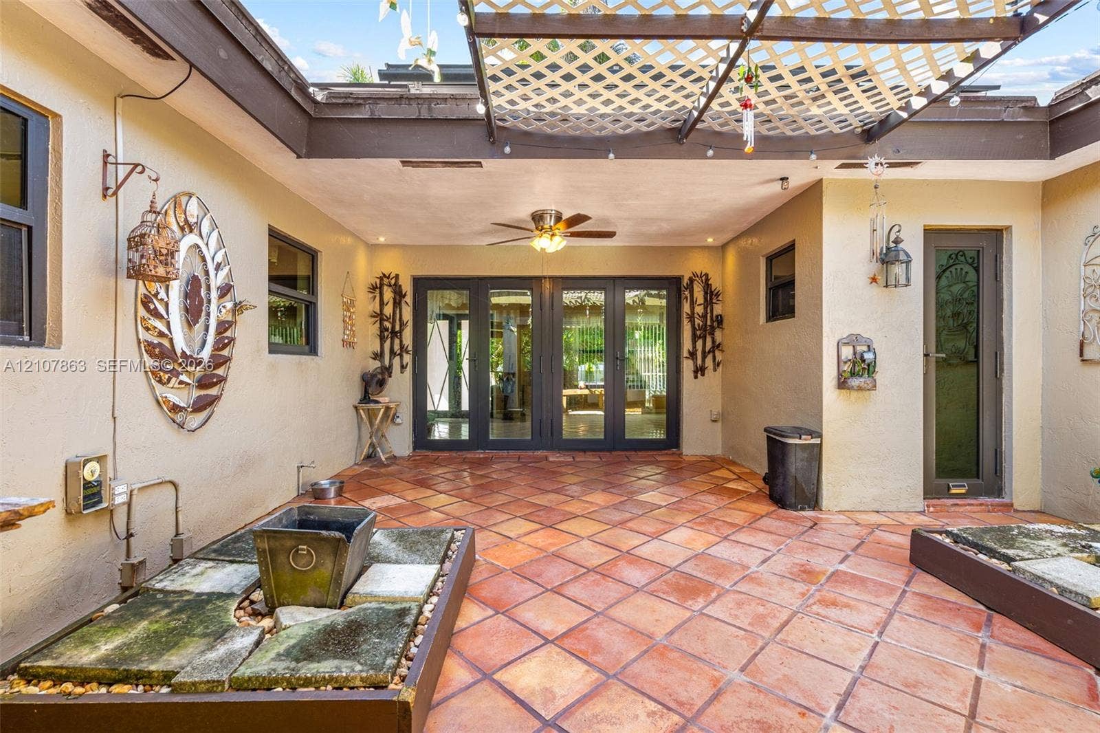 Enclosed courtyard featuring terracotta tile flooring, a structured wooden lattice overhead, multiple built-in stone planters, and a ceiling-mounted fan