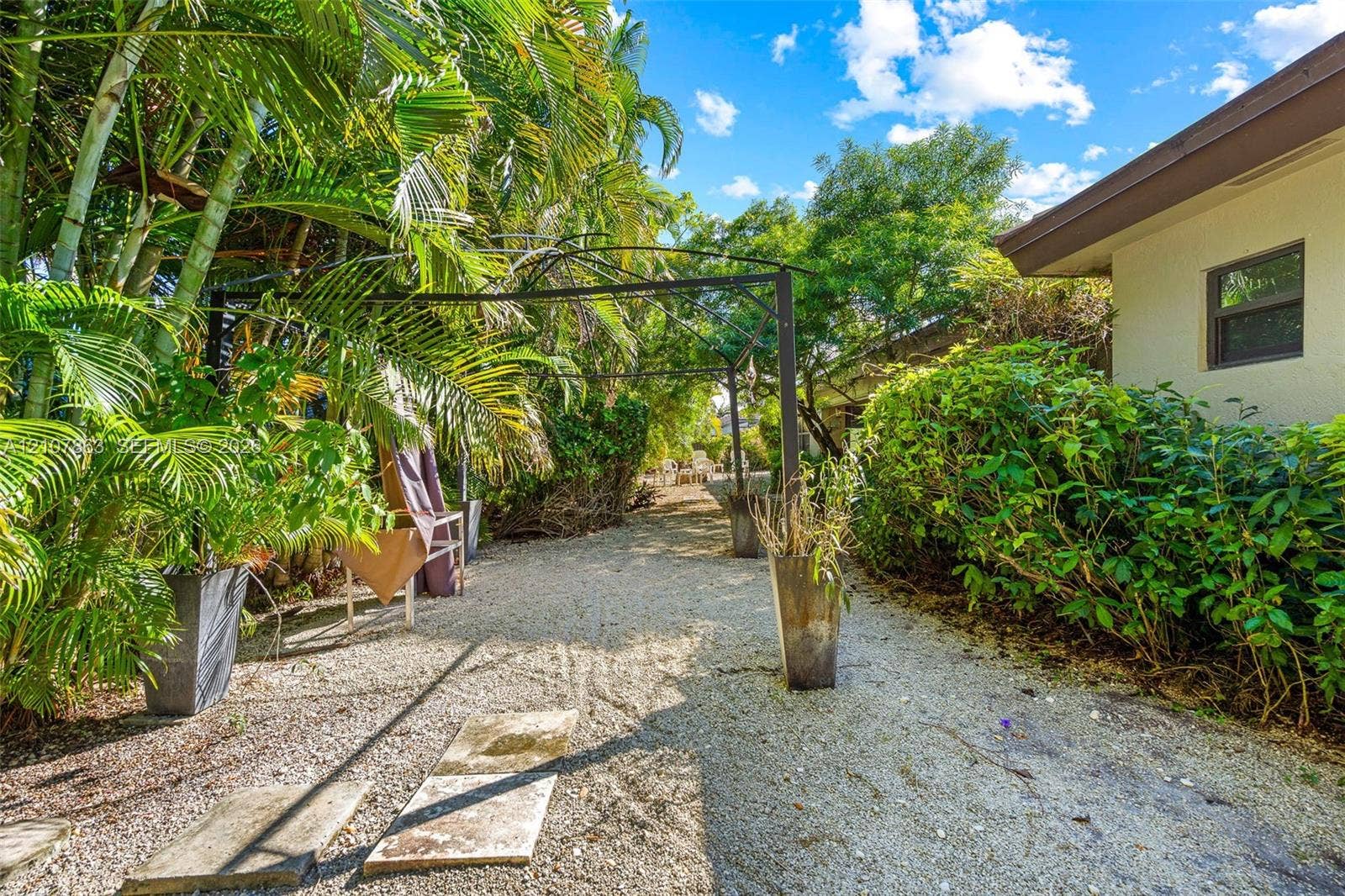 Exterior side area featuring a gravel path, mature palm trees, metal trellis structure, and dense shrubbery
