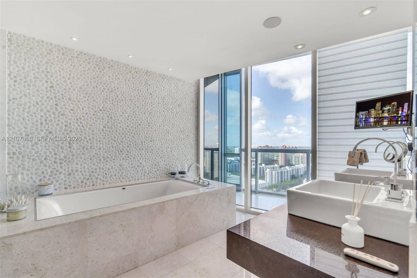 17121 Collins Avenue, #2608, Sunny Isles Beach, FL 33160 Drop-in bathtub with stone-finish surround, textured pebble-style accent wall, dual rectangular vessel sinks, polished stone countertop, and floor-to-ceiling windows