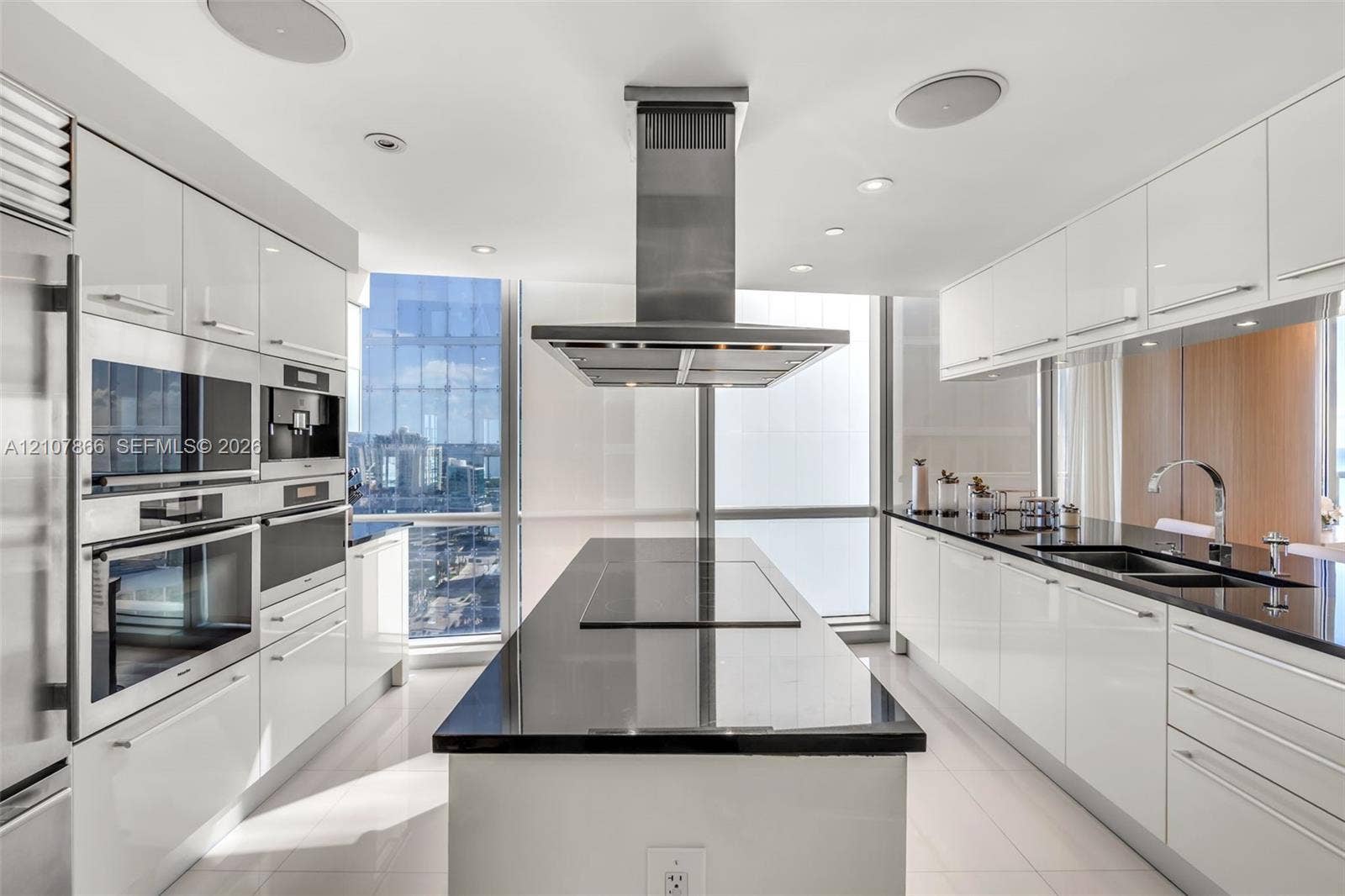 17121 Collins Avenue, #2608, Sunny Isles Beach, FL 33160 Modern kitchen featuring high-gloss white cabinetry, a center island with a dark solid-surface countertop, stainless steel integrated wall ovens, a ceiling-mounted stainless steel vent hood, and floor-to-ceiling windows