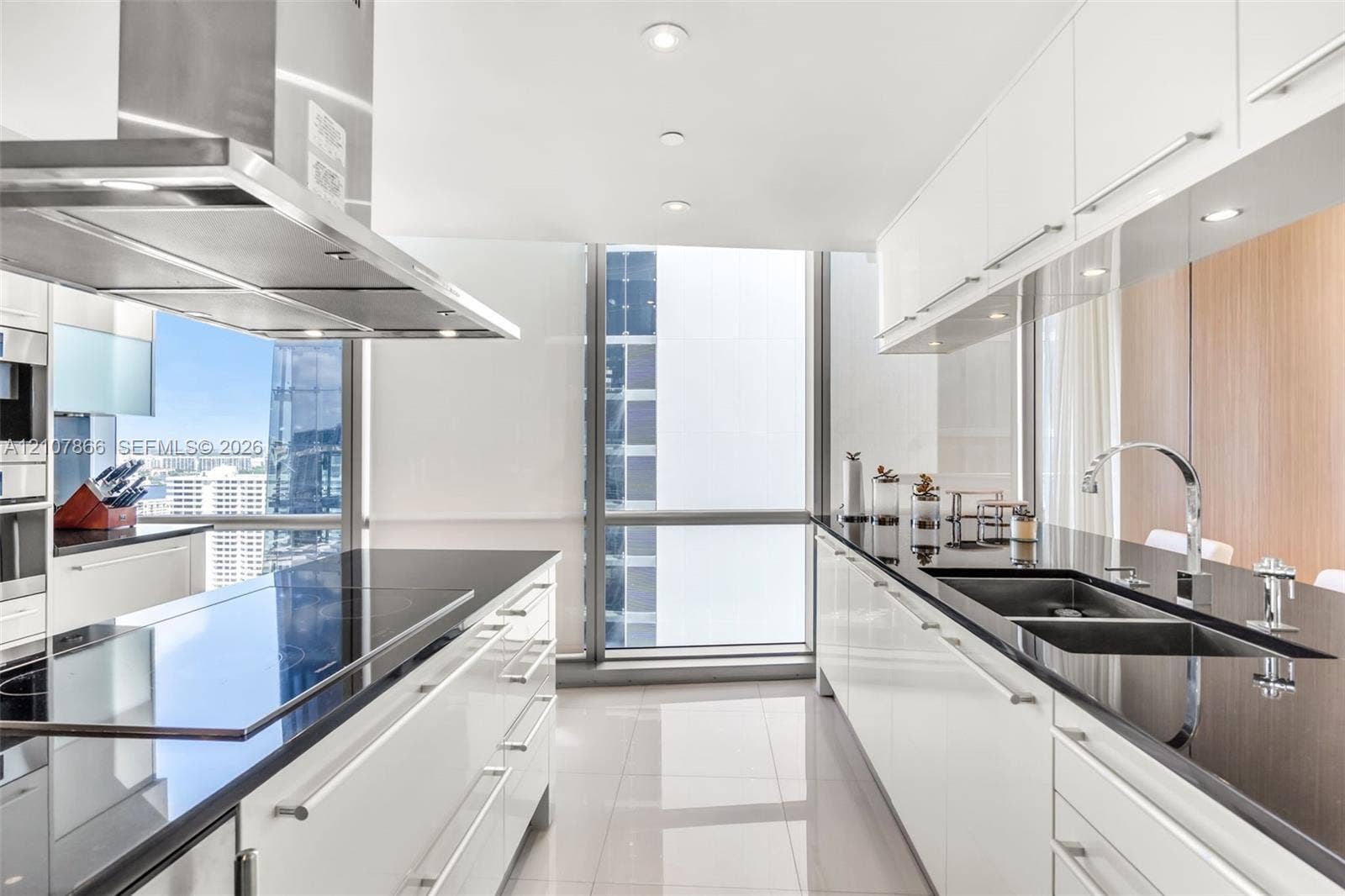 17121 Collins Avenue, #2608, Sunny Isles Beach, FL 33160 Contemporary kitchen featuring flat-panel white cabinetry, contrasting black stone countertops, an integrated stainless steel range hood, double under-mount sinks with a gooseneck faucet, and floor-to-ceiling glass windows