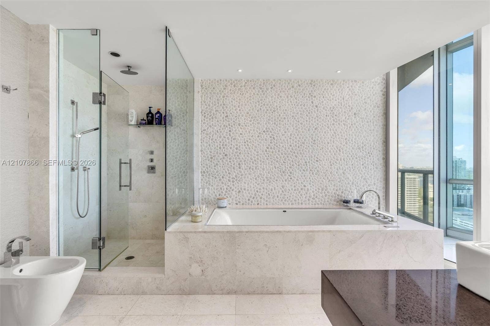 17121 Collins Avenue, #2608, Sunny Isles Beach, FL 33160 Bathroom featuring a built-in soaking tub, glass-enclosed walk-in shower with a rainfall showerhead, stone-tile flooring, textured stone-accent wall, and floor-to-ceiling windows
