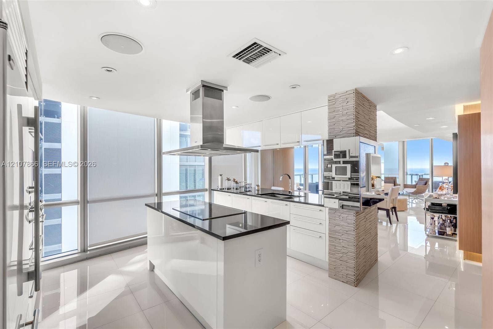 17121 Collins Avenue, #2608, Sunny Isles Beach, FL 33160 Contemporary kitchen featuring a center island with a sleek cooktop, stainless steel chimney-style range hood, high-gloss cabinetry, floor-to-ceiling windows, and a decorative stone-clad pillar