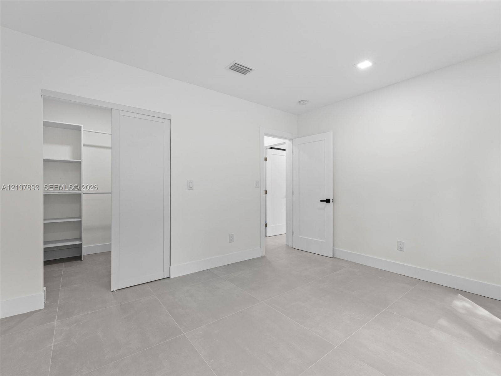 610 NE 164th Terrace, Miami, FL 33162 Interior space featuring large-format grey tile flooring, white walls, recessed ceiling lighting, a walk-in closet with built-in shelving, and a solid-core door with matte black hardware