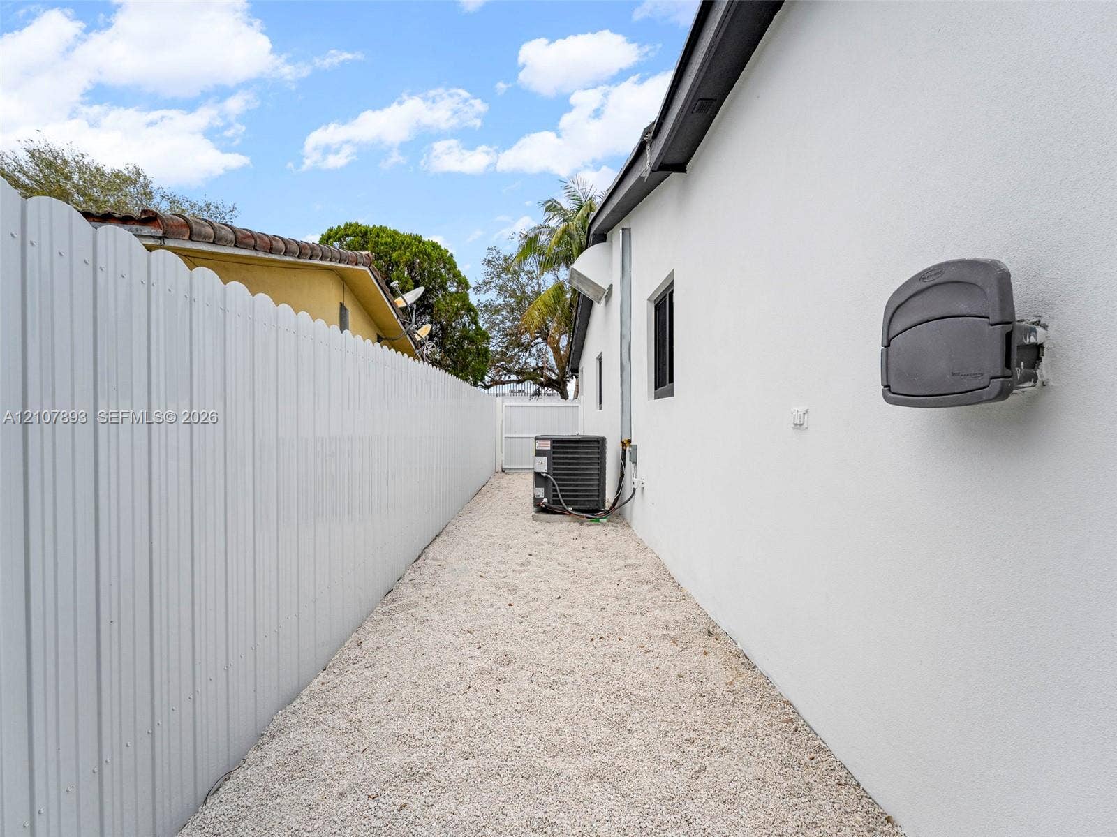 610 NE 164th Terrace, Miami, FL 33162 Exterior side yard featuring white stucco siding, full-height white vinyl fencing, gravel ground cover, and a dedicated HVAC unit