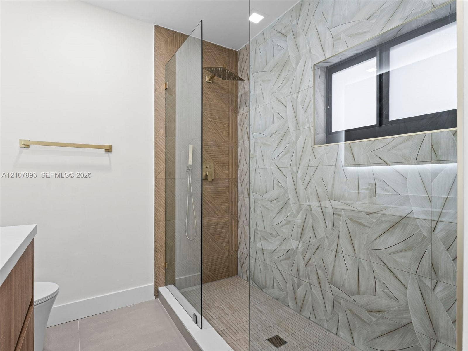610 NE 164th Terrace, Miami, FL 33162 Walk-in shower featuring gold-tone rainfall and handheld showerheads, glass enclosure, leaf-patterned wall tile, and built-in niche lighting